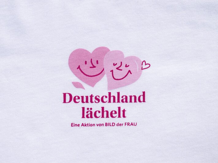 Nahaufnahme eines Logos mit lächelnden Herzen und der Aufschrift "Deutschland lächelt" auf weißem Stoff. | © Bild der Frau/ Ulrike Schacht