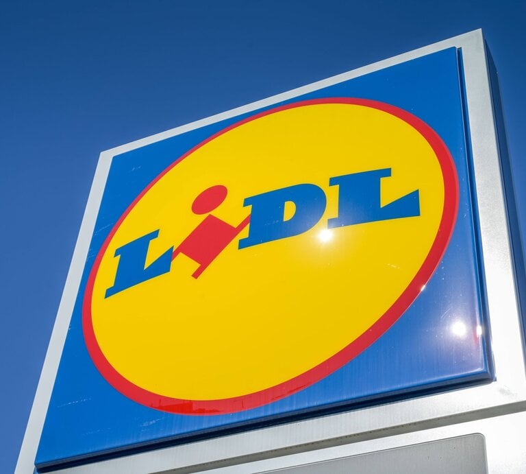 Sonnenstrahlen spiegeln sich auf einem scharf fokussierten Lidl-Logo unter blauem Himmel. | © IMAGO / Schöning