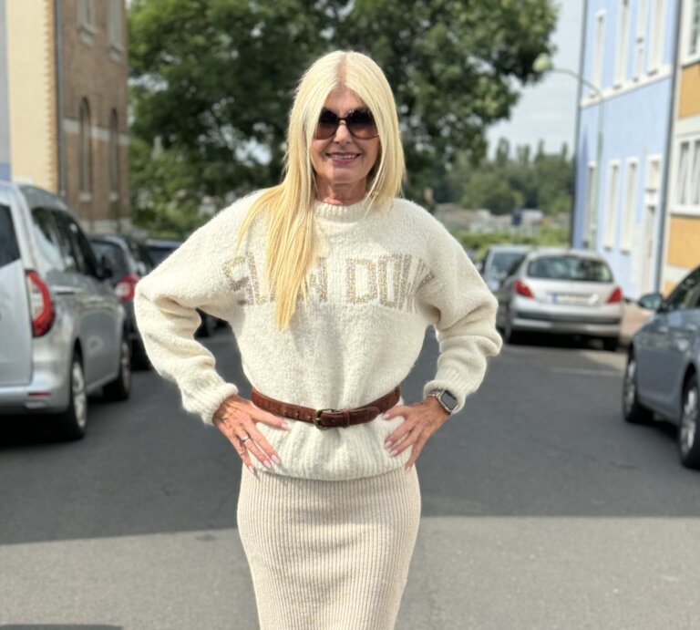 Frau mittleren Alters in einem urbanen Straßenszenario, trägt einen cremefarbenen Strickpullover mit der Aufschrift "LEAD DUKE", dazu passenden langen Strickrock und braune Stiefel.  | © Renate Zott