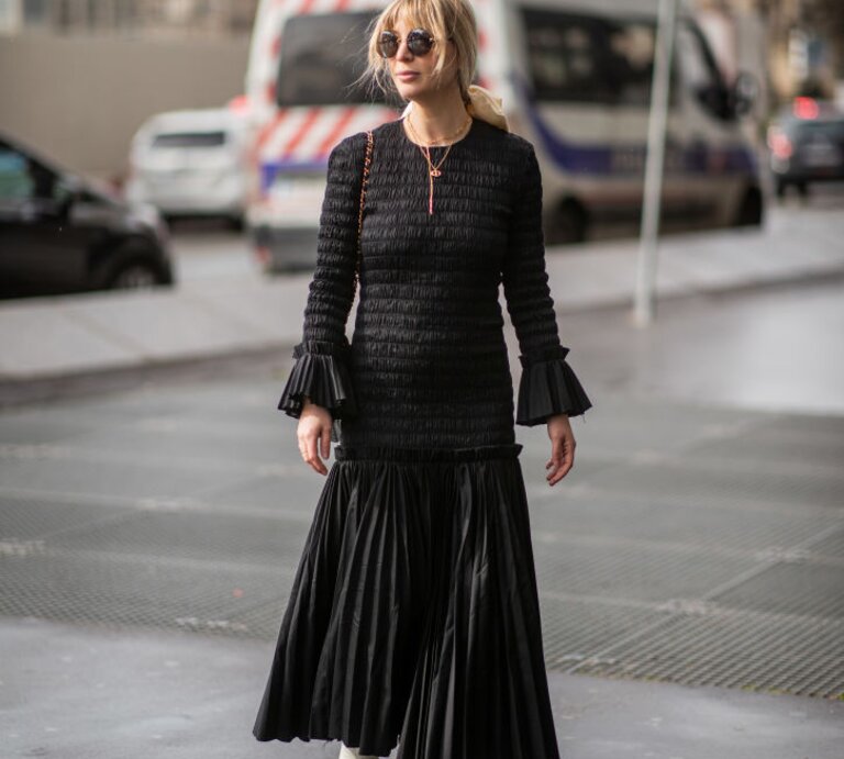 Eine blonde Frau in einem langen, schwarzen Kleid und weißen Stiefeletten spaziert auf einer nassen Straße in der Stadt. | © GettyImages/Christian Vierig 