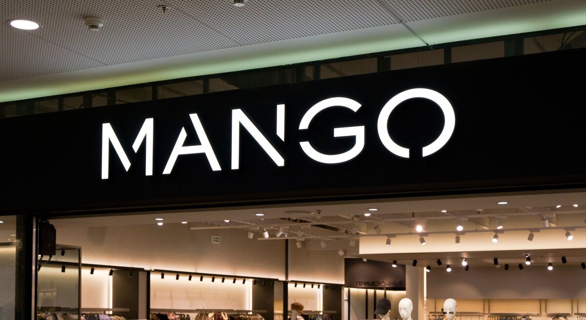 Eingang eines Mango-Geschäfts mit leuchtendem Logo, dahinter moderne Kleidungsausstellung und Schaufensterpuppen.
