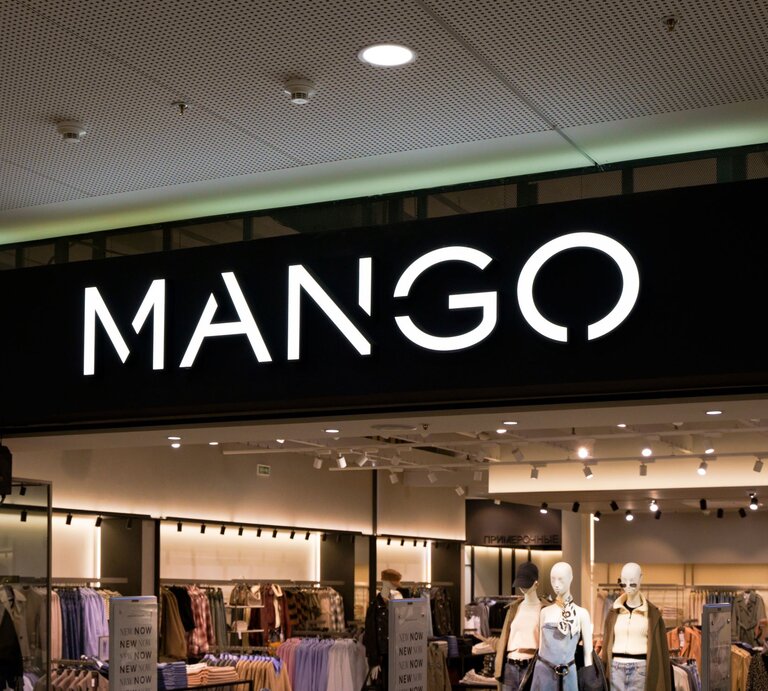Eingang eines Mango-Geschäfts mit leuchtendem Logo, dahinter moderne Kleidungsausstellung und Schaufensterpuppen. | © Adobe Stock/ uladzimirzuyeu