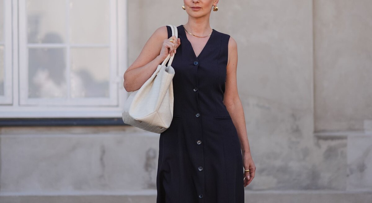 Frau in schwarzem Kleid und Sonnenbrille trägt weiße Tasche, steht vor einem hellen Fenster und einer beige Wand.