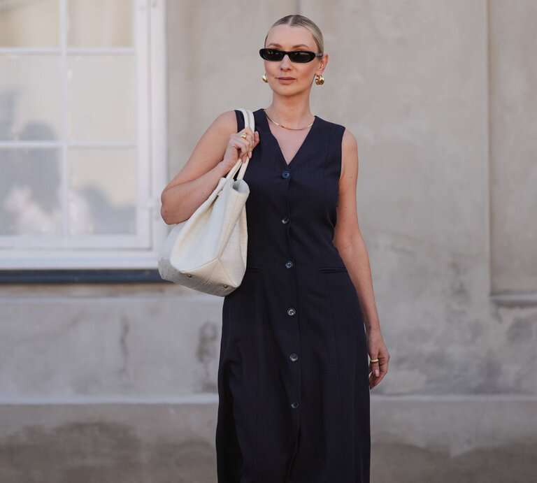 Frau in schwarzem Kleid und Sonnenbrille trägt weiße Tasche, steht vor einem hellen Fenster und einer beige Wand. | © Edward Berthelot/Getty Images