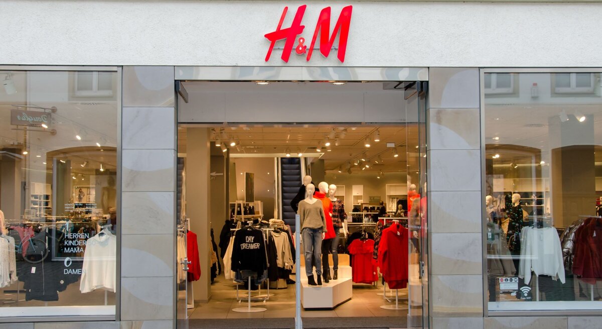 Eingang eines H&M-Geschäfts mit offenen Glastüren. Über dem Eingang befindet sich das rot-weiße H&M-Logo. Im Schaufenster sind Mannequins in aktueller Mode bekleidet, darunter ein rotes Oberteil und eine schwarze Jacke. Schilder im Inneren deuten auf verschiedene Bekleidungsabteilungen wie Herren, Kinder, Mama und Damen hin.