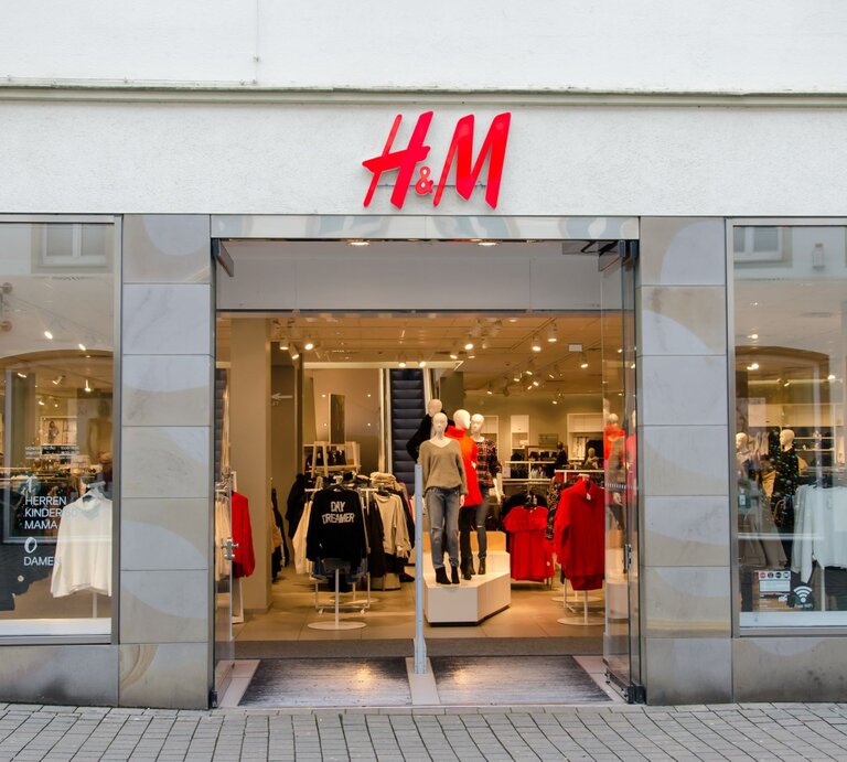 Eingang eines H&M-Geschäfts mit offenen Glastüren. Über dem Eingang befindet sich das rot-weiße H&M-Logo. Im Schaufenster sind Mannequins in aktueller Mode bekleidet, darunter ein rotes Oberteil und eine schwarze Jacke. Schilder im Inneren deuten auf verschiedene Bekleidungsabteilungen wie Herren, Kinder, Mama und Damen hin. | © Adobe Stock/ Олександр Луценко