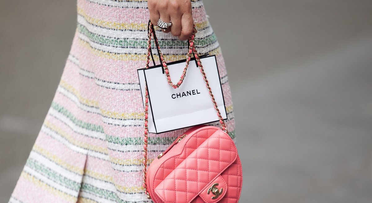 Frau in pastellfarbenem gestreiftem Rock hält eine rosa Chanel-Tasche und eine Chanel-Papiertüte.