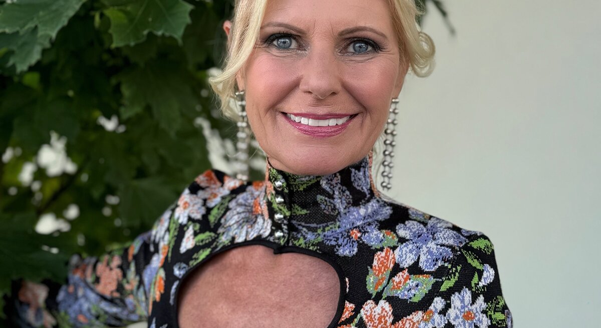 Ältere Frau mit einem strahlenden Lächeln, die in die Kamera blickt. Sie hat blondes Haar und trägt ein farbenfrohes Kleid mit einem floralen Muster und einem auffälligen Ausschnitt am Dekolleté.