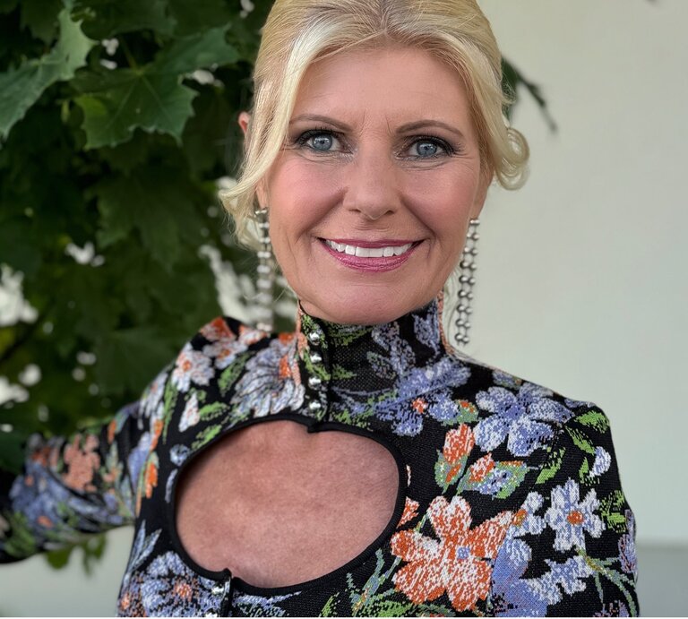 Ältere Frau mit einem strahlenden Lächeln, die in die Kamera blickt. Sie hat blondes Haar und trägt ein farbenfrohes Kleid mit einem floralen Muster und einem auffälligen Ausschnitt am Dekolleté. | © Renate Zott