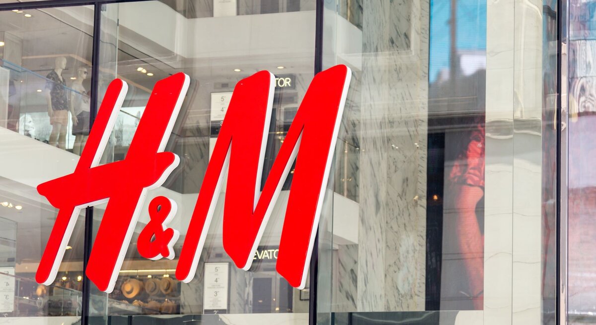 Das Bild zeigt das große, leuchtend rote Logo von H&M an der Fassade eines Geschäfts. Durch die großen Fenster hinter dem Logo sind Mannequins und Teile der Inneneinrichtung des Ladens zu erkennen.