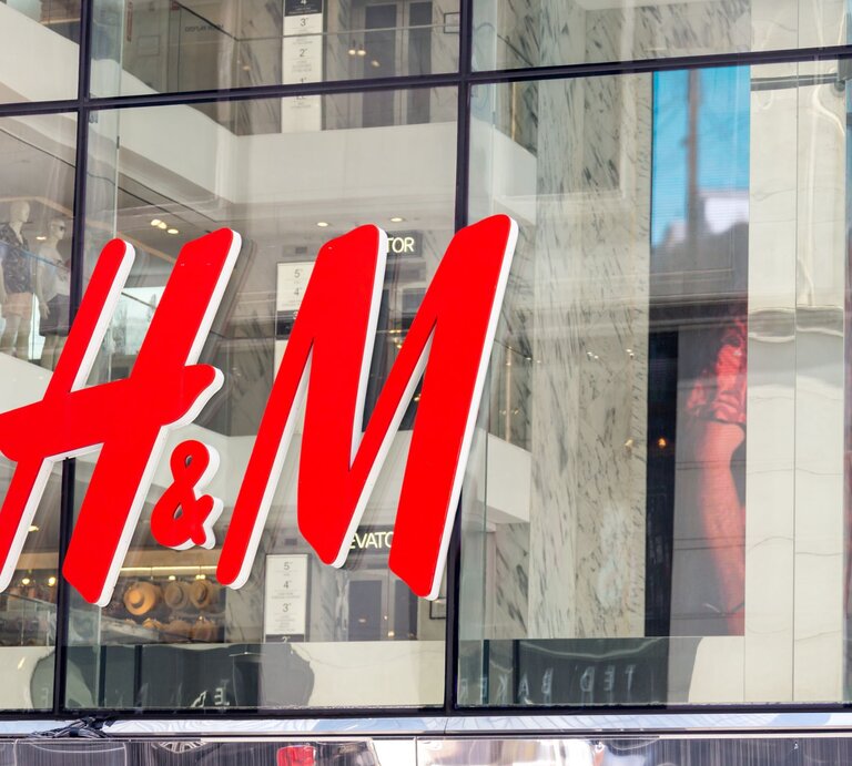 Das Bild zeigt das große, leuchtend rote Logo von H&M an der Fassade eines Geschäfts. Durch die großen Fenster hinter dem Logo sind Mannequins und Teile der Inneneinrichtung des Ladens zu erkennen. | © Adobe Stock/ Mariakray