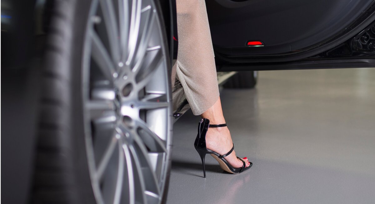 Frau steigt aus einem Auto aus. Sie trägt elegante, glänzende schwarze Stiletto-Sandalen und eine beige Hose. Man sieht nur ein Bein.