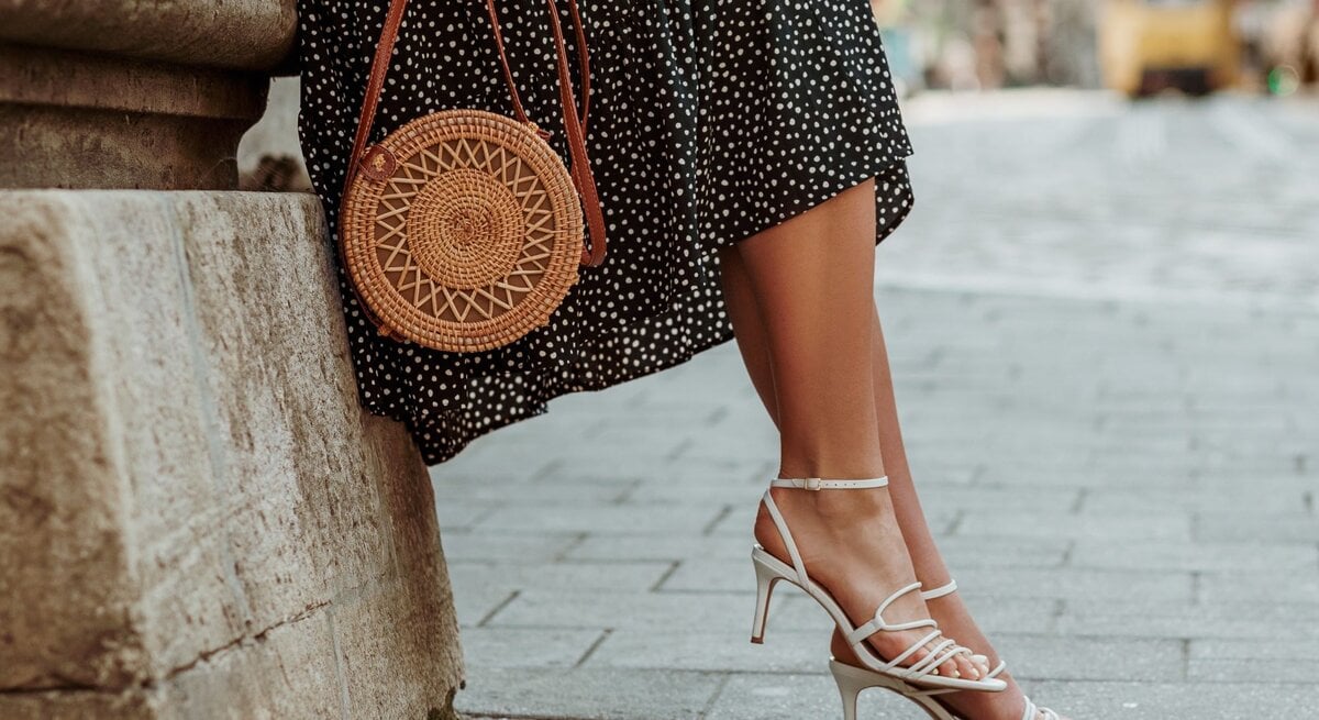 Eine Frau in einem gepunkteten Kleid lehnt an einer Mauer, trägt weiße High Heels und hält eine runde Rattantasche.