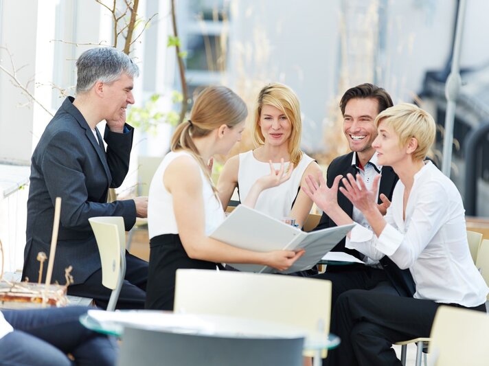 Geschäftsleute, die bei einem informellen Meeting im Freien sitzen. Die Personen sind in Business-Kleidung gekleidet und scheinen in einer lebhaften Diskussion engagiert zu sein. | © Shutterstock / Robert Kneschke