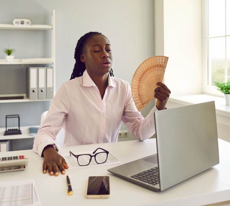 Geschäftsfrau im weißen Hemd kühlt sich im Büro mit einem Fächer, während sie am Laptop arbeitet. | © Adobe Stock/ Studio Romantic