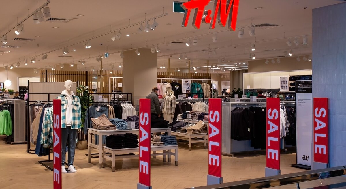 Ein H&M-Bekleidungsgeschäft mit gut beleuchtetem, geräumigem Innenraum. Verschiedene Kleidungsstücke sind auf Mannequins und in Regalen ausgestellt. Auffällige rote "SALE"-Schilder sind im Vordergrund zu sehen, die auf Sonderangebote hinweisen. Im Hintergrund ist das charakteristische H&M-Logo in roten Buchstaben deutlich sichtbar. Der Laden scheint ruhig, ohne viele Kunden, was eine entspannte Einkaufsatmosphäre schafft.