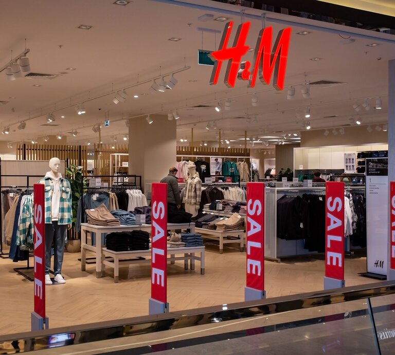 Ein H&M-Bekleidungsgeschäft mit gut beleuchtetem, geräumigem Innenraum. Verschiedene Kleidungsstücke sind auf Mannequins und in Regalen ausgestellt. Auffällige rote "SALE"-Schilder sind im Vordergrund zu sehen, die auf Sonderangebote hinweisen. Im Hintergrund ist das charakteristische H&M-Logo in roten Buchstaben deutlich sichtbar. Der Laden scheint ruhig, ohne viele Kunden, was eine entspannte Einkaufsatmosphäre schafft. | © Adobe Stock/ 8th