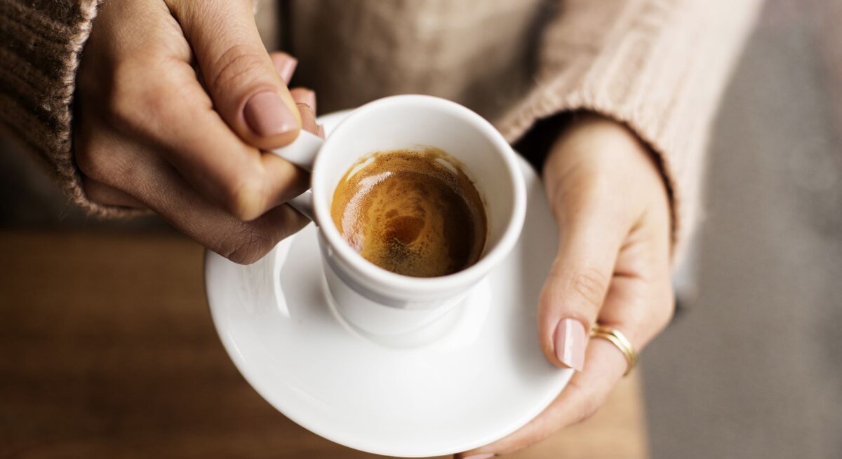 Nahaufnahme von zwei Händen mit gepflegten Nägeln, die eine weiße Espressotasse auf einem Holztisch halten.