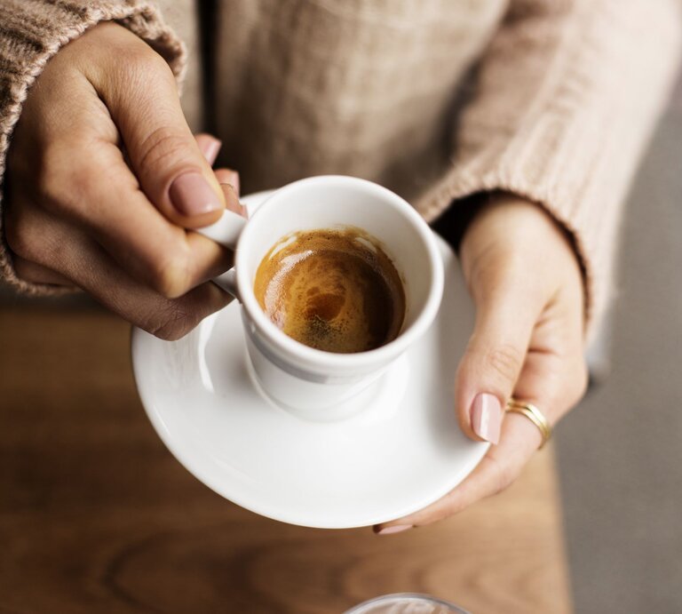 Nahaufnahme von zwei Händen mit gepflegten Nägeln, die eine weiße Espressotasse auf einem Holztisch halten. | © Getty Images/ Pinkybird
