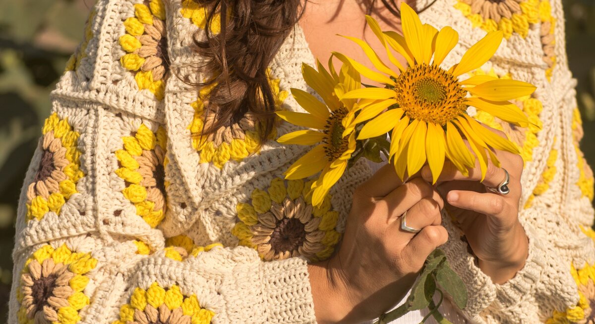 Nahaufnahme einer Frau in einem gestrickten Sonnenblumenpullover, die zwei große gelbe Sonnenblumen in den Händen hält.