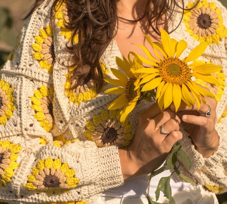 Nahaufnahme einer Frau in einem gestrickten Sonnenblumenpullover, die zwei große gelbe Sonnenblumen in den Händen hält. | © Adobe Stock/ marisol