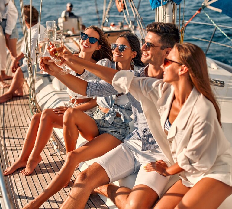 Lächelnde Männer und Frauen in Sommerkleidung stoßen auf einem Segelboot bei sonnigem Wetter an. | © Adobe Stock/ Vasyl