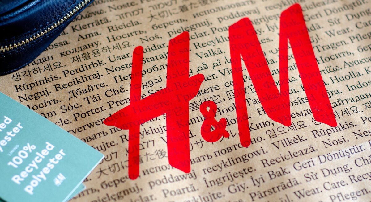 Zu sehen ist eine braune Tüte, auf der in verschiedenen Sprachen "Tragen. Pflegen. Wiederverwenden" zu lesen ist sowie in großen roten Buchstaben "H&M". Auf der linken Seite liegt ein hellgrünes Etikett mit der Aufschrift "100% Recycled Polyester" und ein Stück eines dunkelblauen Portemonnaies.