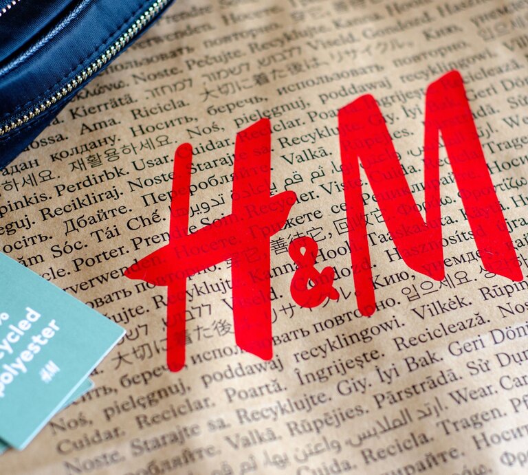 Zu sehen ist eine braune Tüte, auf der in verschiedenen Sprachen "Tragen. Pflegen. Wiederverwenden" zu lesen ist sowie in großen roten Buchstaben "H&M". Auf der linken Seite liegt ein hellgrünes Etikett mit der Aufschrift "100% Recycled Polyester" und ein Stück eines dunkelblauen Portemonnaies. | © Adobe Stock/ Олександр Луценко
