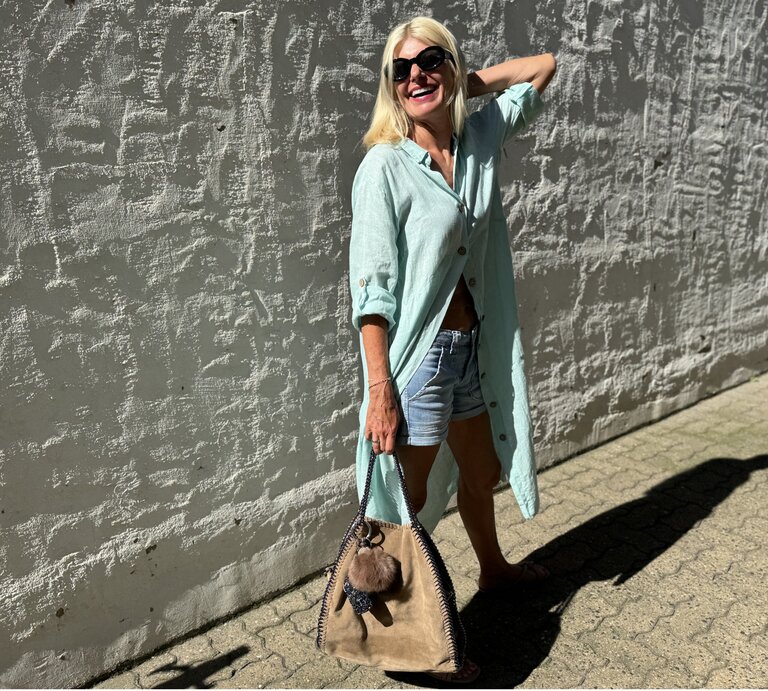 Frau mit blonden Haaren und Sonnenbrille, die sich leger an einer grauen Wand im Freien anlehnt. Sie trägt ein hellblaues, offenes Hemd über einem weißen Top und blaue Denim-Shorts. Ihr Outfit wird durch eine große beige Handtasche mit Fransen abgerundet. | © Renate Zott