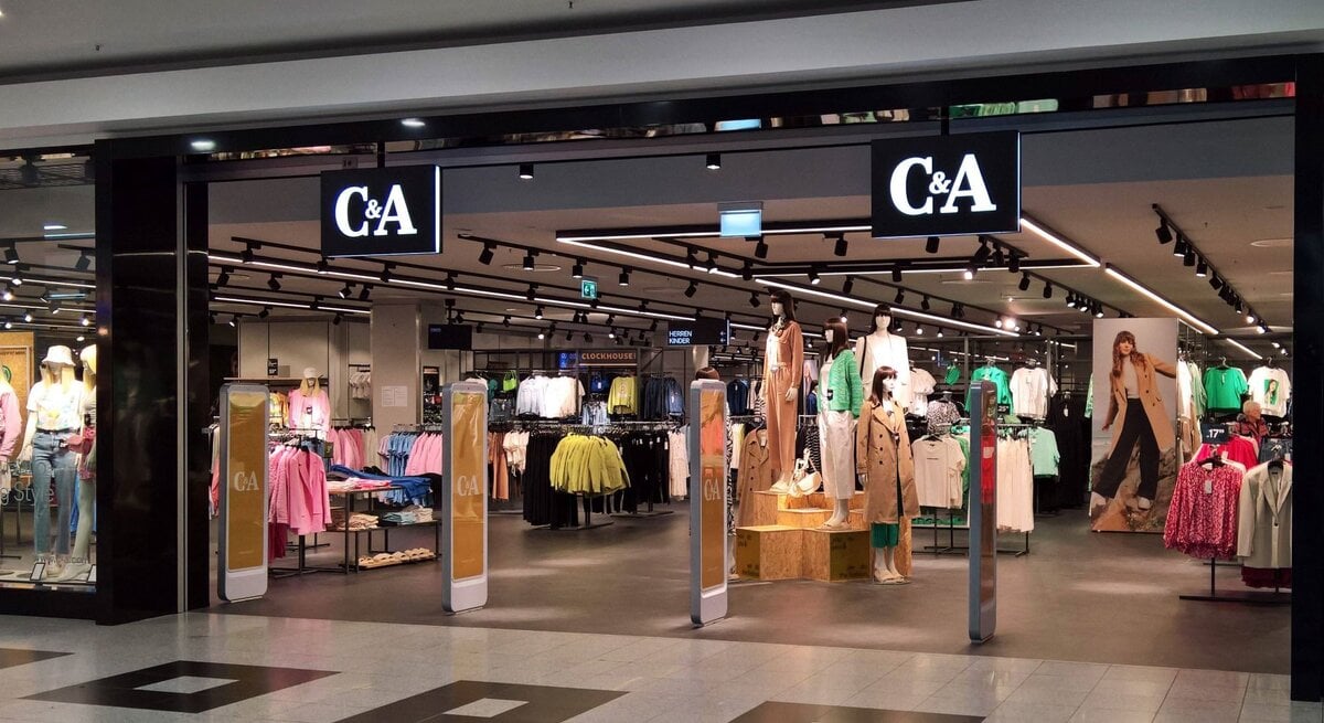 Der Eingangsbereich einer C&A-Filiale in einem Einkaufszentrum. Über dem Eingang hängt ein großes, beleuchtetes C&A-Logo. Im Inneren des Geschäfts sind mehrere Kleidungsständer und Schaufensterpuppen zu sehen, die aktuelle Mode präsentieren. Die Schaufensterpuppen tragen verschiedene Outfits, von Freizeitkleidung bis zu eleganteren Stücken. An der rechten Seite des Eingangs ist ein großes Werbeplakat mit einem Model in einem trendigen Outfit. Der Boden des Einkaufszentrums ist mit dunklen und hellen Fliesen gemustert, und die Beleuchtung im Laden wirkt einladend.