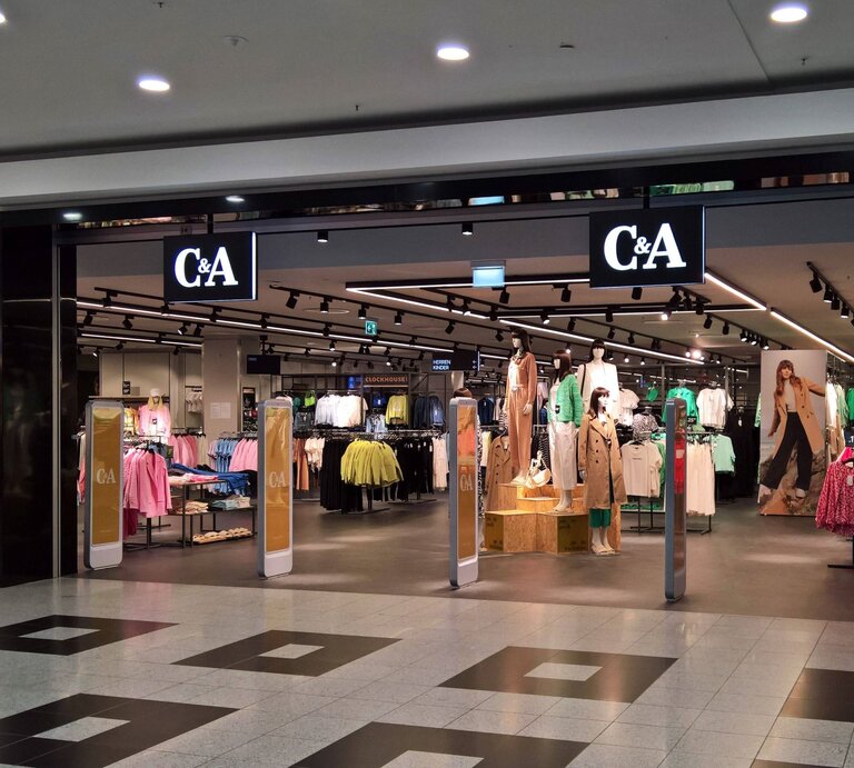 Der Eingangsbereich einer C&A-Filiale in einem Einkaufszentrum. Über dem Eingang hängt ein großes, beleuchtetes C&A-Logo. Im Inneren des Geschäfts sind mehrere Kleidungsständer und Schaufensterpuppen zu sehen, die aktuelle Mode präsentieren. Die Schaufensterpuppen tragen verschiedene Outfits, von Freizeitkleidung bis zu eleganteren Stücken. An der rechten Seite des Eingangs ist ein großes Werbeplakat mit einem Model in einem trendigen Outfit. Der Boden des Einkaufszentrums ist mit dunklen und hellen Fliesen gemustert, und die Beleuchtung im Laden wirkt einladend. | © IMAGO / Manfred Segerer