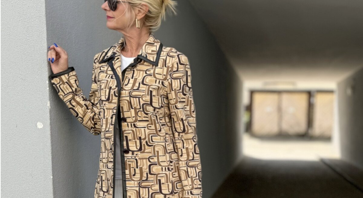 Frau mit blonden Haaren, die zu einem lockeren Knoten hochgesteckt sind. Sie trägt eine Sonnenbrille und eine gemusterte Jacke in Beige- und Brauntönen mit geometrischen Mustern. 