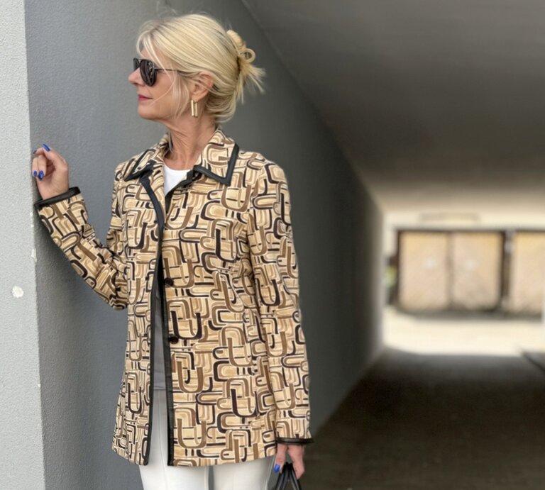 Frau mit blonden Haaren, die zu einem lockeren Knoten hochgesteckt sind. Sie trägt eine Sonnenbrille und eine gemusterte Jacke in Beige- und Brauntönen mit geometrischen Mustern.  | © Renate Zott