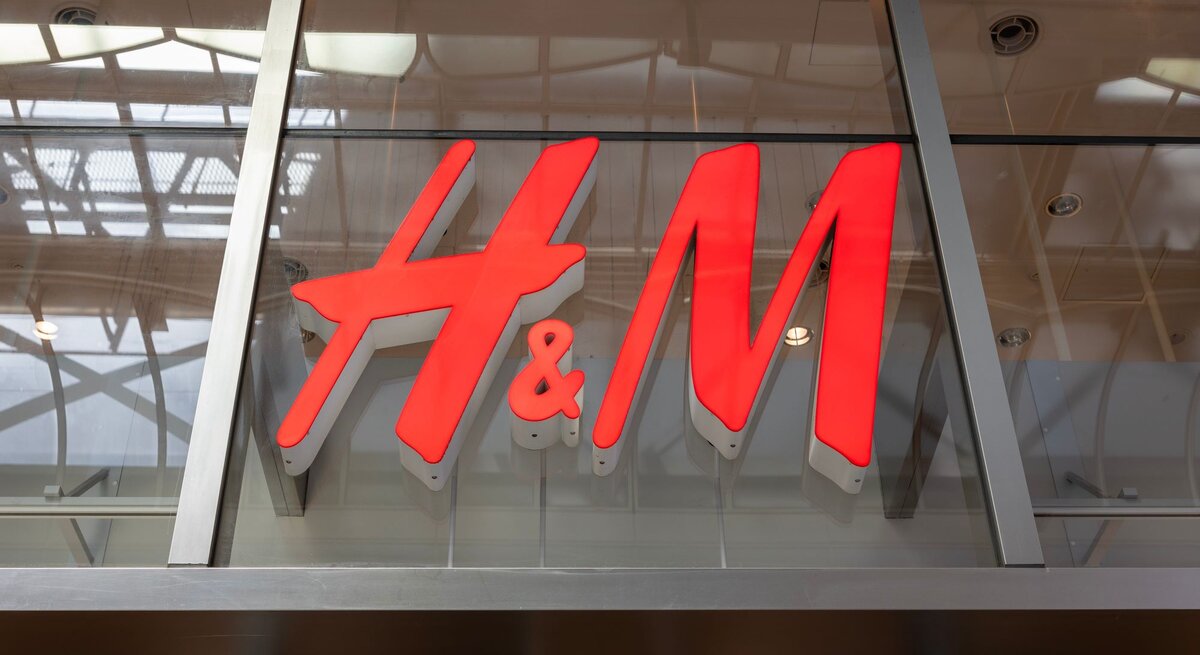 Großes H&M-Logo in leuchtendem Rot an einer gläsernen Fassade, reflektiert das Innenlicht.