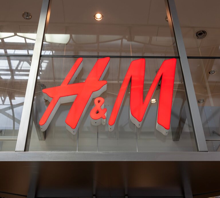 Großes H&M-Logo in leuchtendem Rot an einer gläsernen Fassade, reflektiert das Innenlicht. | © Adobe Stock/ J_News_photo