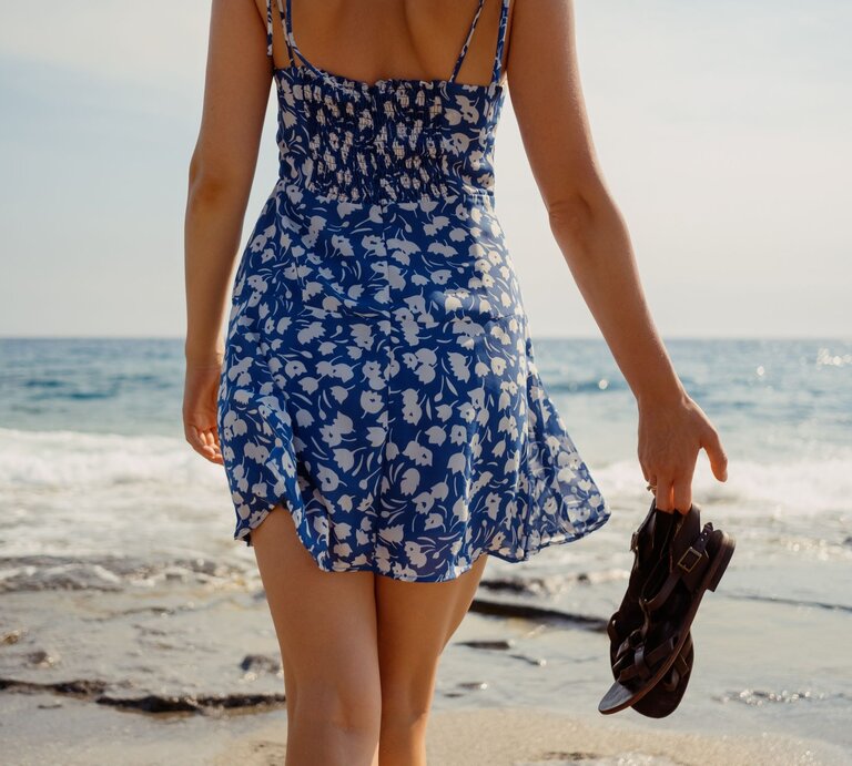 Frau in geblümtem Kleid spaziert barfuß mit Sandalen in der Hand an der Küste entlang bei ruhigem Meer. | © Adobe Stock/ arthurhidden