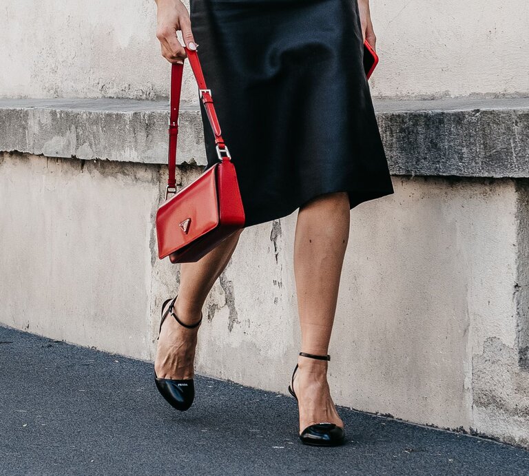 Frau in schwarzem Midirock geht auf Gehweg, trägt rote Handtasche und schwarze Sandalen mit Riemchen. | © Adobe Stock/ photo-lime