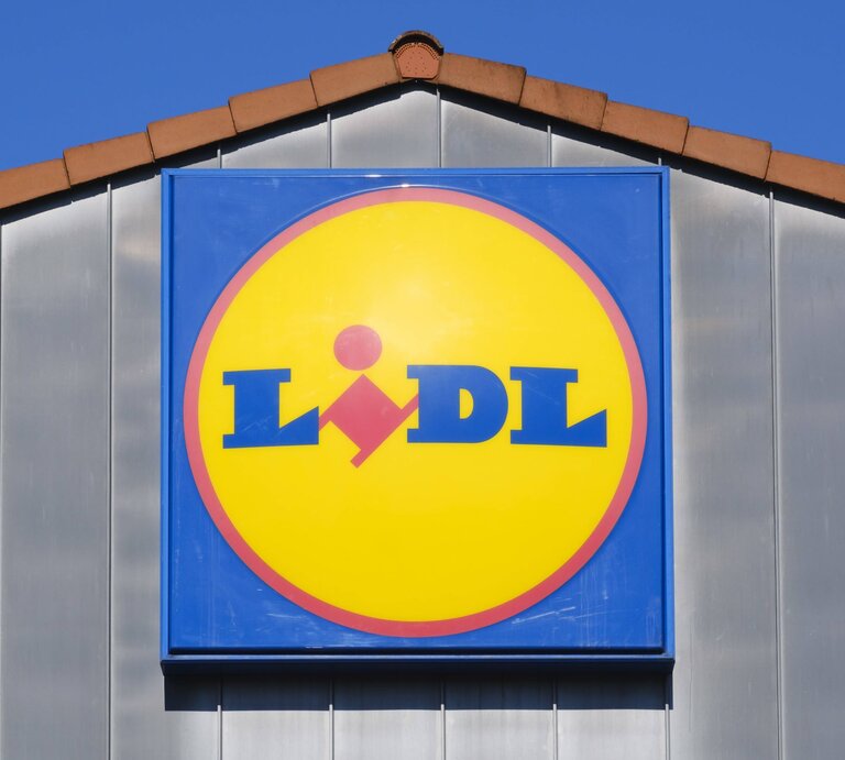 Leuchtendes Lidl-Logo auf blauem Schild, montiert an einer Fassade unter blauem Himmel. | © IMAGO / CHROMORANGE