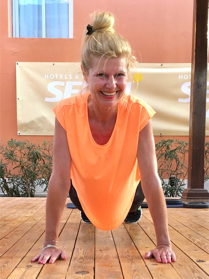 Frau mittleren Alters, die in einem leuchtend orangefarbenen ärmellosen Oberteil auf dem Boden eine Übung macht, vermutlich eine Art Liegestütz oder Plank. | © Renate Zott
