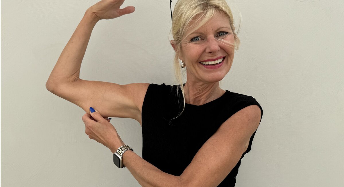 Frau in einem ärmellosen, schwarzen Oberteil. Sie hebt einen Arm und zeigt mit der anderen Hand auf ihren Oberarm, was ihre trainierte und fitte Erscheinung betont. Sie trägt eine Uhr und hat blaue Fingernägel.