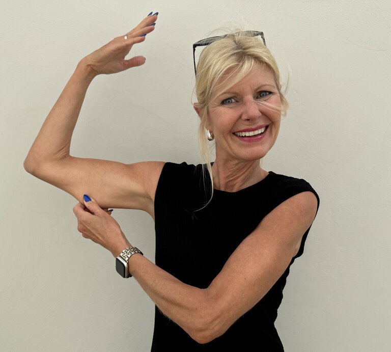 Frau in einem ärmellosen, schwarzen Oberteil. Sie hebt einen Arm und zeigt mit der anderen Hand auf ihren Oberarm, was ihre trainierte und fitte Erscheinung betont. Sie trägt eine Uhr und hat blaue Fingernägel. | © Renate Zott