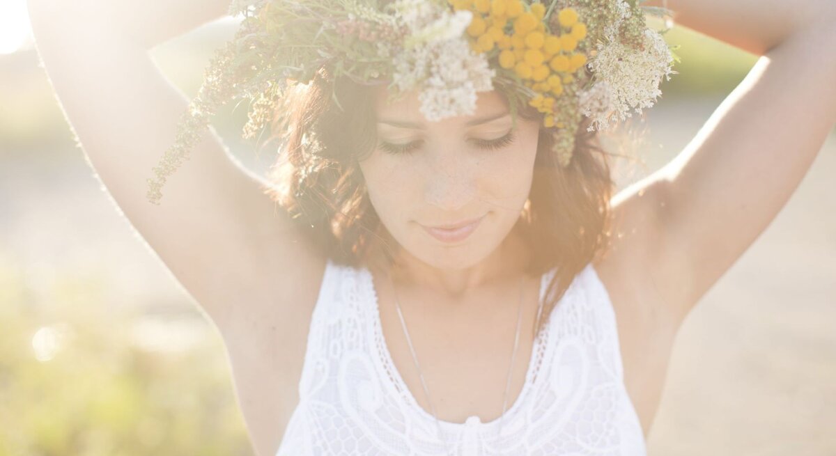 Eine Frau mit Blumenkranz und weißem Spitzenkleid steht verträumt im Sonnenlicht mit erhobenen Armen.