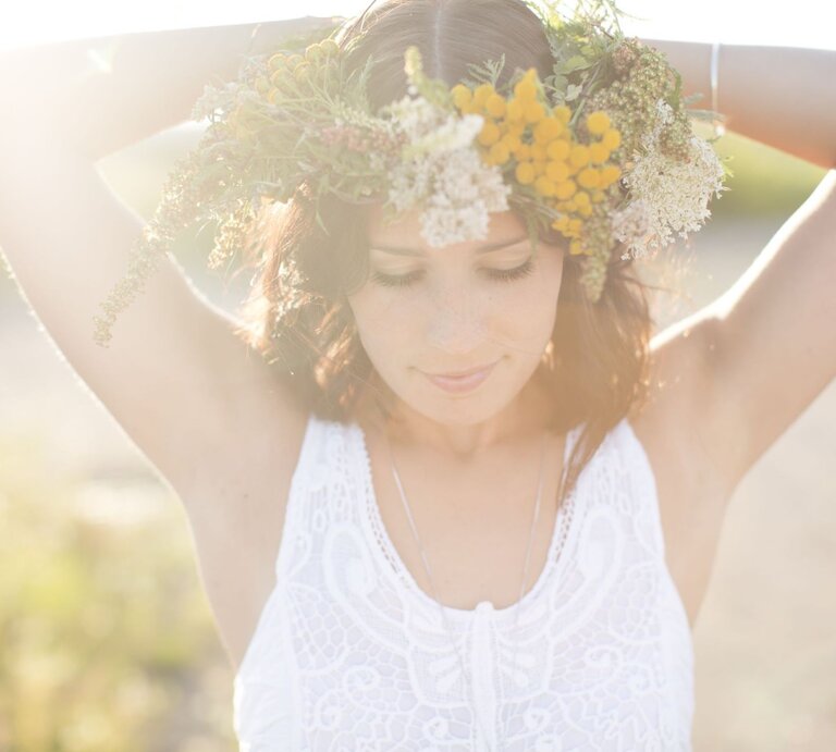 Eine Frau mit Blumenkranz und weißem Spitzenkleid steht verträumt im Sonnenlicht mit erhobenen Armen. | © Getty Images/ Johner Images