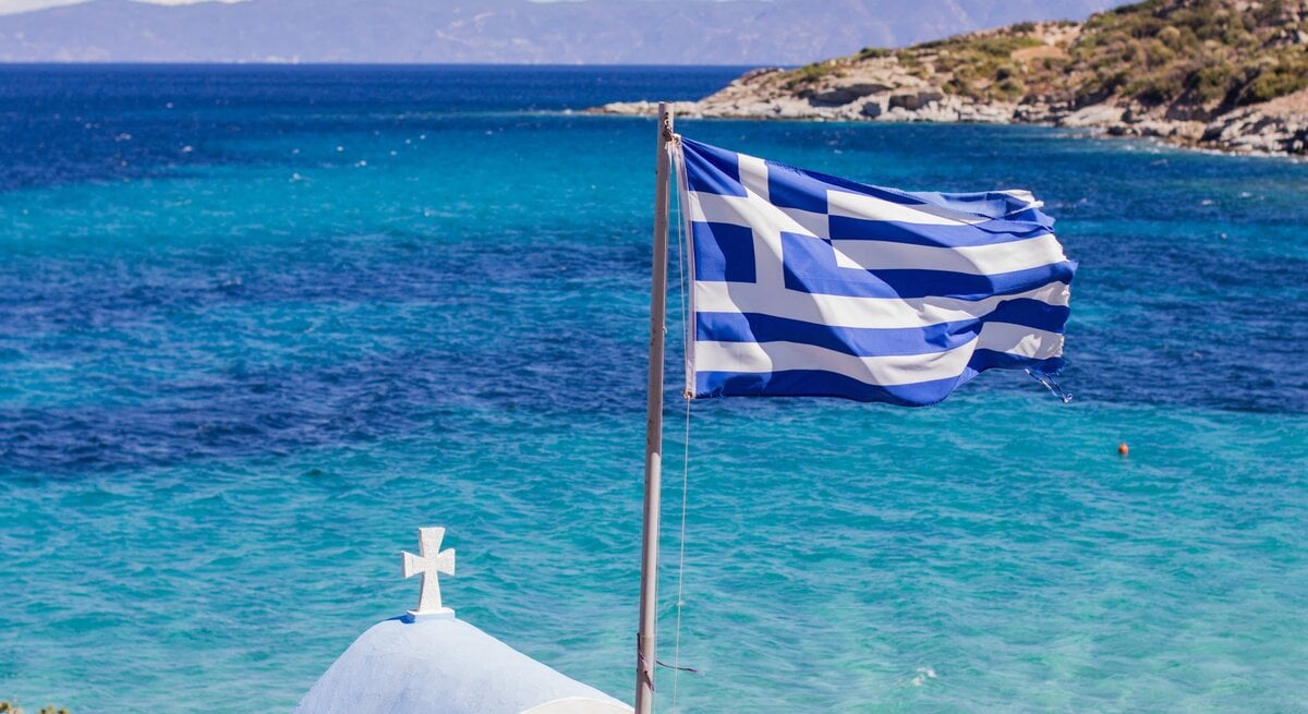 Symbolbild für Luxus-Sandalen: Die griechische Flagge weht im Wind vor türkisblauem Meer, dahinter eine kleine weiße Kapelle mit Kreuz und felsiger Küste.
