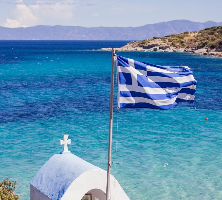 Symbolbild für Luxus-Sandalen: Die griechische Flagge weht im Wind vor türkisblauem Meer, dahinter eine kleine weiße Kapelle mit Kreuz und felsiger Küste. | © Adobe Stock/ mitarart
