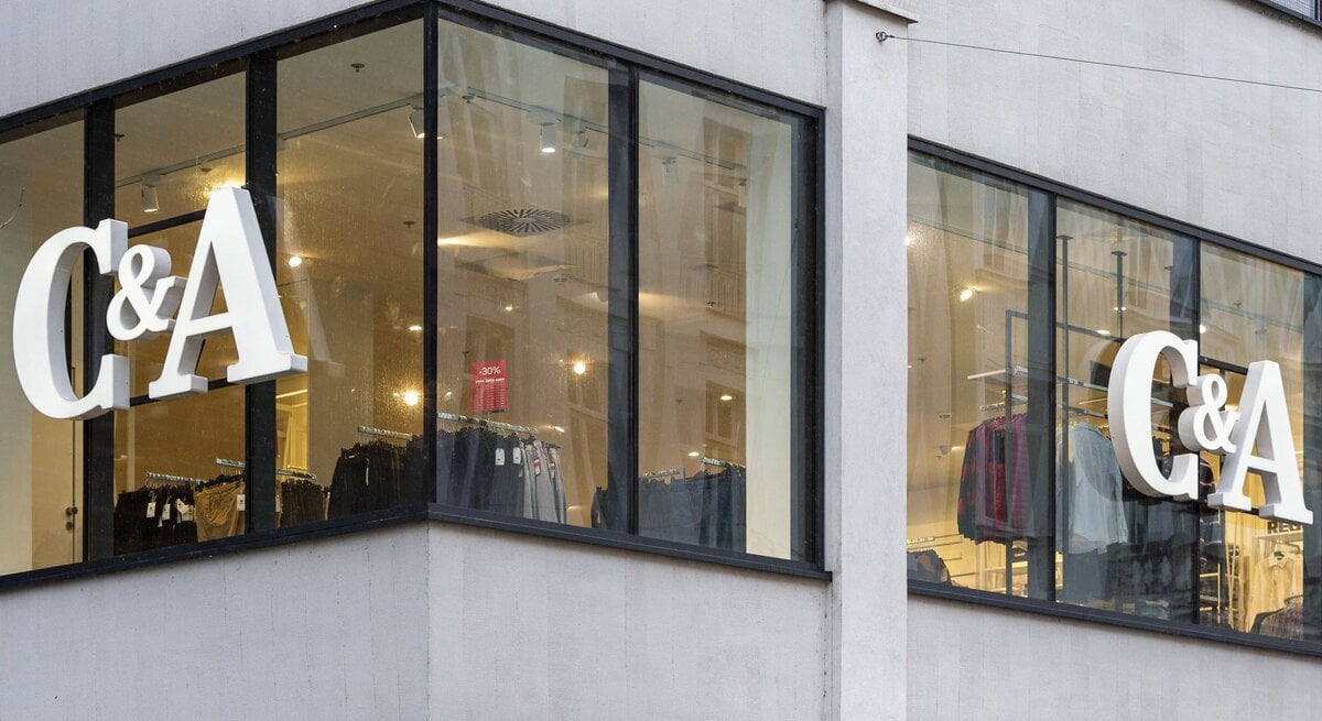  Ein Foto der Eckfassade eines modernen Gebäudes mit großen Fenstern, in denen zwei identische "C&A"-Logos in weißen Lettern zu sehen sind. Hinter dem Glas sind Kleidungsstücke aufgehängt, was darauf hindeutet, dass es sich um ein Bekleidungsgeschäft handelt. Es scheint ein bewölkter Tag zu sein, und das Licht im Inneren des Ladens lässt die Logos und die angebotene Ware hell erscheinen. Ein Rabattschild mit "-30%" ist in der Auslage sichtbar.