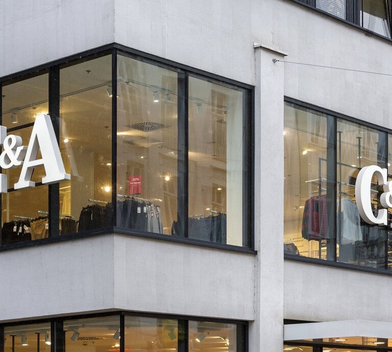  Ein Foto der Eckfassade eines modernen Gebäudes mit großen Fenstern, in denen zwei identische "C&A"-Logos in weißen Lettern zu sehen sind. Hinter dem Glas sind Kleidungsstücke aufgehängt, was darauf hindeutet, dass es sich um ein Bekleidungsgeschäft handelt. Es scheint ein bewölkter Tag zu sein, und das Licht im Inneren des Ladens lässt die Logos und die angebotene Ware hell erscheinen. Ein Rabattschild mit "-30%" ist in der Auslage sichtbar. | © IMAGO / CHROMORANGE