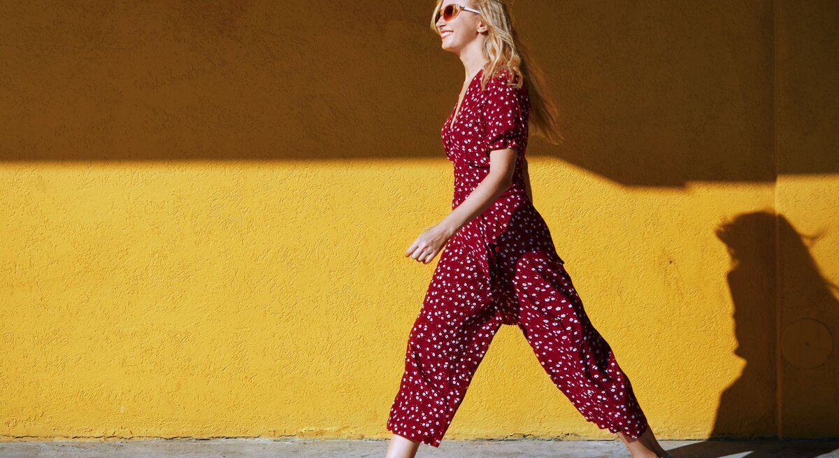 Stilvolle blonde Dame in rot-weiß gepunktetem Jumpsuit und blauen Sneakern läuft lächelnd an einer gelben Wand entlang.