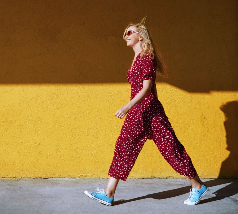 Stilvolle blonde Dame in rot-weiß gepunktetem Jumpsuit und blauen Sneakern läuft lächelnd an einer gelben Wand entlang. | © Getty Images/ Arman Zhenikeyev