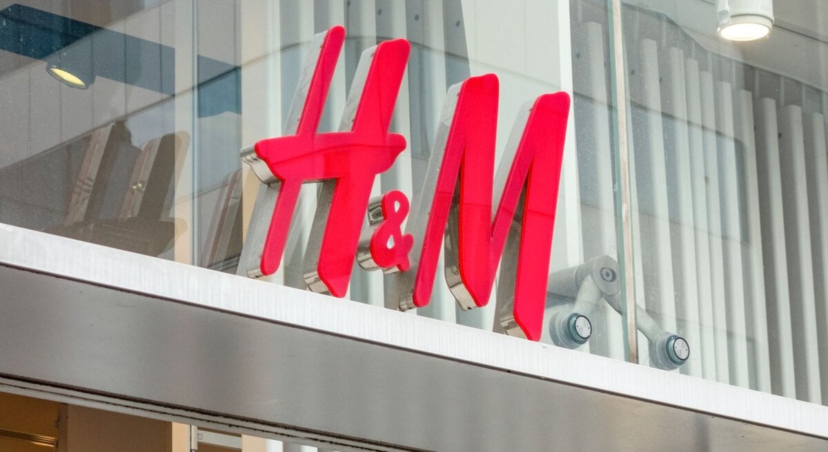 Das Bild zeigt das leuchtend rote Logo von H&M, das an einer Ladenfront befestigt ist. Das Logo ist vor einem gläsernen Hintergrund mit Sicht auf innenliegende Strukturen und Gegenstände platziert. Darüber sind moderne Beleuchtungselemente angebracht.